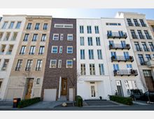 Straßenansicht mit mehreren modernen Wohngebäuden in unterschiedlichen Fassadenstilen, darunter Klinker-, Putz- und Steinfassaden. Die Gebäude haben große Fenster, Balkone mit Metallgeländern und vereinzelt dekorative Elemente, mit angrenzenden kleinen Vorgärten.