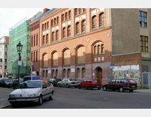 Straßenszene mit Fassade eines historischen Backsteingebäudes im Stil der Industriearchitektur, gekennzeichnet durch hohe Rundbogenfenster und symmetrische Details. Links ist ein mit grünem Baugerüst eingerüstetes Gebäude zu sehen, während mehrere geparkte Autos die Straße säumen.