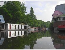 Ein ruhiger Kanal mit Hausbooten am Ufer, umgeben von dichten Bäumen und vereinzelten Gebäuden, darunter ein rundes Gebäude mit einem rosa Dach im rechten Bildbereich.