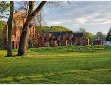 Ruine eines historischen Backsteingebäudes mit gotischen Bögen, umgeben von einer grünen Wiese und Bäumen; im Hintergrund ein Fachwerkgebäude und bewaldete Hügel.
