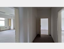 Leerer Flur mit weißen Wänden und Teppichboden, der zu mehreren offenen Türen führt. Durch eine Tür ist ein heller Raum mit großen Fenstern und Heizkörpern sichtbar.