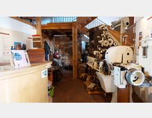 Innenansicht eines Museums mit einer Ausstellung zur Tabakverarbeitung. Zu sehen sind eine große, historische Tabakverarbeitungsmaschine, eine sitzende Schaufensterpuppe in Arbeitskleidung sowie Infotafeln und historische Gegenstände wie Säcke und Modelle.