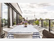 Moderne Dachterrasse mit Glasgeländern, einem großen Esstisch mit weiß-blau gestreifter Tischdecke und geflochtenen Stühlen, dekoriert mit Pflanzen und Blumen; im Hintergrund ist der Berliner Fernsehturm sichtbar.