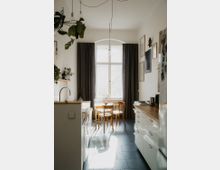 Helle Küche mit einer Kombination aus weißer und Holzoptik. Ein Holztisch mit vier Stühlen steht vor einem großen Fenster mit dunklen Vorhängen, umgeben von schlichten weißen Wänden und minimalistischem Dekor.