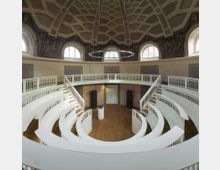 Runder historischer Vorlesungssaal mit mehreren weißen, konzentrischen Sitzreihen und umlaufendem Geländer. Eine Kuppeldecke mit geometrischem Muster sowie dekorative Wandmalereien und große Fenster bestimmen den Raum.