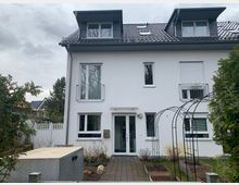 Zweistöckiges Wohnhaus mit weißer Fassade und grauem Satteldach, ausgestattet mit Dachgauben. Vor dem Gebäude befinden sich ein gepflasterter Weg, Hochbeete und bogenförmige Metallrankgerüste. Im Hintergrund sind Bäume und Nachbarhäuser zu sehen.