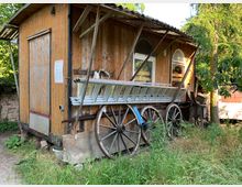 Holzschuppen mit gelblich-brauner Fassade, kleinen Fenstern und Satteldach, umgeben von Gras und Bäumen. Alte Holzwagenräder und gelagerte Leitern sind an der Außenwand befestigt.