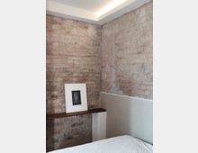Ecke eines Raumes mit unverputzten Ziegelwänden, einer schmalen Holzablage mit einem gerahmten Kunstbild und einem weißen Bett im Vordergrund. Die Beleuchtung erfolgt durch eine eingelassene Deckenleuchte.