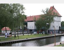 Historisches Schleusenbauwerk mit grauem Mauerwerk und roten Ziegeldächern, umgeben von Bäumen und Grünflächen. Im Vordergrund fließt ein Kanal mit befestigtem Ufer und Anlegestellen, während ein Hinweisschild neben der Schleuse platziert ist.