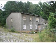 Ein verlassenes, zweistöckiges Gebäude mit grauem Putz und zugemauerten Fenstern, umgeben von dichtem Wald und überwuchertem Pflasterweg. Im Vordergrund wachsen Gräser und Sträucher wild.