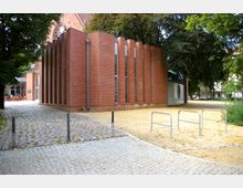 Backsteingebäude mit vertikal angeordneten Fenstern und modernen architektonischen Elementen, umgeben von Bäumen. Im Vordergrund ein gepflasterter Weg und ein Schotterbereich mit Fahrradständern.