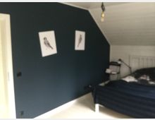 Ein Schlafzimmer mit einer schrägen weißen Decke und einer dunkelblauen Wand, an der zwei Bilder mit Vogelzeichnungen hängen. Im Raum stehen ein Bett mit dunkler Bettwäsche und ein kleiner Tisch mit einer Lampe und Büchern.