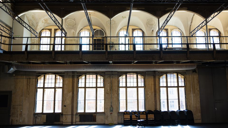 Große, hohe Industriehalle mit zwei Ebenen, hohen Rundbogenfenstern und einer umlaufenden Stahl-Metallgalerie unter der Decke. Sichtbare Trägerkonstruktionen und eine gealterte Putzwand prägen den Raum, am Boden stehen gestapelte Stühle vor den Fenstern.