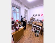 Ein kleiner Raum mit Holzboden und weißen Wänden, ausgestattet mit einem großen Fenster sowie mehreren Arbeitsflächen. Auf den Tischen und Regalen befinden sich diverse Gegenstände, darunter Schneiderpuppen, Bastelmaterialien und Dekorationsartikel, was auf eine kreative Arbeitsumgebung hinweist.