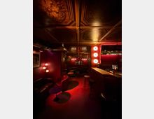 Innenraum einer stilvollen Bar/Lounge mit rot gedämpfter Beleuchtung, gepolsterten Sitzbänken und kleinen Tischen sowie einem langen Tresen mit Barhockern. Auffällig sind die dekorierte Kassettendecke, runde Wandlampen und ein spiegelnder Bereich im Hintergrund.