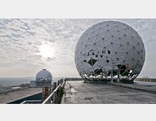 Zwei große, kugelförmige Radarkuppeln auf einer Betonplattform mit Graffiti, umgeben von einer weitläufigen Waldlandschaft unter einem bewölkten Himmel. Die größere Kuppel steht im Vordergrund, während die kleinere im Hintergrund sichtbar ist.