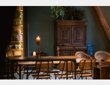 Wohnraum mit rustikalem Charme: Im Vordergrund stehen ein Holztisch und geflochtene Sitzmöbel, im Hintergrund ein antiker Holzschrank und ein Regal mit Pflanzen und Dekorobjekten, von warmem Kerzenlicht beleuchtet. Die Wand ist in dunklem Grün gestrichen, und neben Holzbalken fügt sich die natürliche Einrichtung harmonisch ein.