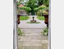 Blick durch eine offene Tür auf einen gepflasterten Hof mit einem runden Springbrunnen in der Mitte. Der Hof ist von symmetrisch gepflanzten, kleinen Bäumen und Blumenbeeten umgeben, im Hintergrund stehen Wohnhäuser mit roten Ziegeldächern und ein grünes Eingangstor.