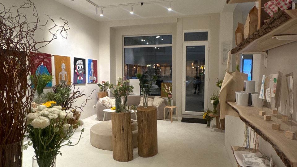 Heller, moderner Laden- oder Galerieraum mit weißem Boden, Tracklights an der Decke und großen Fenstern sowie einer Glastür zum Außenbereich. Im Raum stehen Baumstamm-Tische und Vasen mit Blumen, an der Wand hängen farbige Kunstbilder und rechts befinden sich Regale mit ausliegenden Produkten.