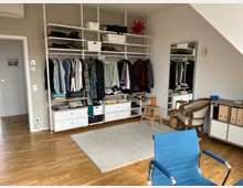 Ein Schlafzimmer oder Ankleidezimmer mit offenem weißen Regalsystem, auf dem Kleider hängen und gefaltet sind, sowie Schubladen und Körben für Stauraum. Im Raum befinden sich ein großer Spiegel, Holzmöbel, ein blauer Stuhl, ein beiger Teppich und ein Parkettboden.