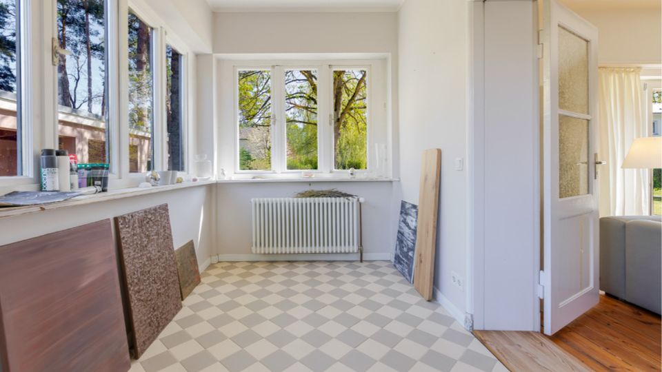 Heller, leerer Innenraum mit großen Fenstern auf zwei Seiten, einer niedrigen Fensterbank und Blick ins Grüne. In der Mitte steht ein Heizkörper unter dem Fenster, der Boden ist mit hellgrau-weißem Rautenmuster gefliest, und rechts führt eine offene Tür in einen angrenzenden Raum mit Holzboden.