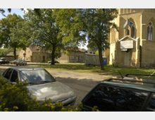 Straßenszene mit parkenden Autos vor einem kleinen Platz mit großen Bäumen. Rechts im Bild ist ein Backsteingebäude mit gotischen Fenstern und einem Portal, das vermutlich eine Kirche ist; dahinter stehen Wohnhäuser.