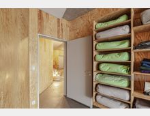 Ein Innenraum mit Holzverkleidung an den Wänden und Regalen, in denen grüne und graue Kissen oder Lagerrollen gestapelt sind. Eine geöffnete Tür führt in einen modernen, minimalistisch gestalteten Raum mit hellen Wänden, geflochtenen Sitzkissen und dekorativen Elementen.