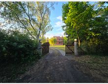Ein ländlicher Weg führt durch ein offenes Holztor zu einem roten Backsteinhaus mit weißen Fensterrahmen, umgeben von einem gepflegten Rasen und hohen Bäumen im Hintergrund. Links und rechts des Tors stehen Säulen mit runden Leuchten, und die Umgebung ist von dichtem grünen Laub geprägt.