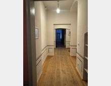 Schmaler, langer Flur mit Holzdielenboden und weißen Wänden, die mit schlichten schwarzen Zierleisten dekoriert sind. Im Hintergrund sind ein Türdurchgang mit Rundbogen und teilweise ein weiterer Raum sichtbar, beleuchtet von einer modernen Hängelampe.