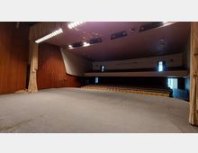 Blick von einer Bühne in einen Theatersaal mit schlichten Holzwänden, herunterhängenden Vorhängen und mehreren Sitzreihen, die sich im Parkett und auf einem Balkon befinden. Die Beleuchtung ist gedämpft, und an der Decke sind mehrere Lichtquellen sichtbar.