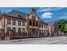 Historisches Gebäude aus roten Ziegelsteinen mit cremefarbenen Fassadenakzenten, Spitzdach und markanter Mittelachse, beschriftet mit „Amtsgericht“. Umgeben von einem metallischen Zaun mit Backsteinsockel, im Vordergrund eine Straße mit Verkehrsinseln.