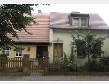 Zweistöckiges Einfamilienhaus mit hellgelber Fassade und rotem Ziegeldach. Vor dem Haus befinden sich ein kleiner Garten, ein Zaun aus Holz und Ziegelpfeilern sowie eine gepflasterte Einfahrt; ein Baum und Sträucher rahmen die Fassade ein.