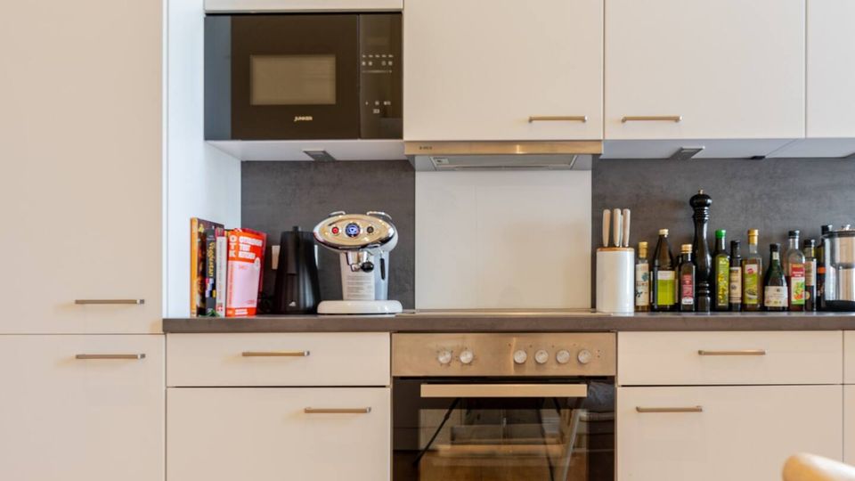 Moderne Einbauküche mit weißen Schrankfronten, dunkler Arbeitsplatte und grauer Rückwand, ausgestattet mit Mikrowelle, Dunstabzug und Backofen. Auf der Arbeitsfläche und auf den Oberschränken stehen mehrere Flaschen, Küchenutensilien und Gläser; davor liegt ein Teppich auf Holzboden.