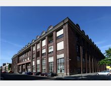 Backsteinfassade eines großen historischen Industriegebäudes mit einer Reihe von Rundbogenfenstern im oberen Bereich und bodentiefen Fenstern im Erdgeschoss. Das Gebäude befindet sich an einer Straßenecke, umgeben von geparkten Autos und vereinzelten Bäumen.