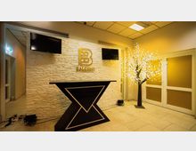 Empfangsbereich mit heller Steinwand, darauf das goldene Logo "Balcony" und zwei schwarze Bildschirme. Im Vordergrund steht ein schwarzer Tresen mit geometrischem Muster, rechts daneben ein dekorativer Baum mit leuchtenden Zweigen.