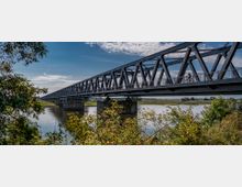 Stählerne Fachwerkbrücke über einen Fluss in einer ländlichen Umgebung, mit Radfahrern auf dem Gehweg der Brücke, umgeben von Bäumen und Sträuchern im Vordergrund. Der Fluss spiegelt den klaren Himmel, während die Landschaft im Hintergrund flach ist.