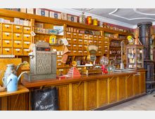 Ein historischer Tante-Emma-Laden mit einer hölzernen Verkaufstheke, darauf eine antike Registrierkasse und Waage. Im Hintergrund sind Holzregale mit beschrifteten Schubladen und alten Warenverpackungen sowie Dekorationsgegenstände zu sehen.
