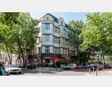 Ecke eines mehrstöckigen Wohnhauses mit cremefarbenen Fassaden und dunklen Horizontalstreifen, im Erdgeschoss eine Kita mit buntem Eingang. Vor dem Gebäude stehen geparkte Autos, und der Bereich ist von großen, grünen Bäumen gesäumt.