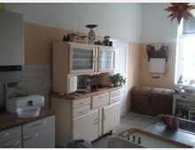 Traditionelle Küche mit cremefarbenem Küchenschrank im Retro-Stil, Holzarbeitsflächen und altem Küchenzubehör. Im Hintergrund stehen eine Holztruhe und ein schmaler Wandregal mit Handtuch, daneben ein Fenster mit Tageslicht.