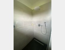 Geflieste Dusche mit hellen Wandfliesen und dunkler Duschwanne, ausgestattet mit großer Regendusche an der Decke und Handbrause an der Wand. In der Ecke verläuft eine Ablageleiste entlang der Wand.