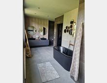 Modernes Badezimmer mit einer freistehenden schwarzen Badewanne, einem Doppelwaschtisch mit runden Wandspiegeln und wandmontierten Dekorationsspiegeln. Der Raum besitzt eine beige-graue Farbpalette mit großen Bodenfliesen und minimalistischer Einrichtung.