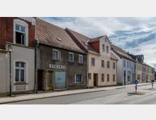 Straßenszene mit einer Reihe von kleinen Wohn- und Geschäftshäusern, darunter eine Bäckerei mit einem schlichten grauen Fassadenfinish und gelbem Eingangsrahmen. Die Gebäude haben rote Ziegeldächer und Fenster im traditionellen Stil, neben einer gepflasterten Straße mit Bürgersteig und Straßenlaternen.