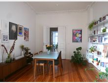 Esszimmer in einer Altbauwohnung mit hohen Decken und Stuckverzierungen. Der Raum enthält einen Holztisch mit Stühlen, eine längliche Kommode, viele Wandbilder, weiße Bücherregale mit Büchern und Pflanzen sowie Holzdielenboden.