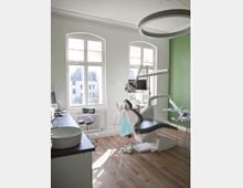 Zahnarztpraxis mit hellem, modernem Behandlungsraum. Im Raum befinden sich ein Zahnarztstuhl, ein Behandlungsgerät, ein Waschbecken auf einer Arbeitsfläche und große Fenster, die Tageslicht hereinlassen; die Wand ist teilweise grün gestrichen.