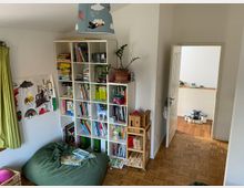 Kinderzimmer mit einem weißen Regal voller Bücher und Spielsachen, einem grünen Sitzsack und bemalten Bildern an der Wand. Der Raum hat Holzboden, eine grüne Gardine, und eine geöffnete Tür führt zu einem angrenzenden Bereich mit Holzspielzeug und Bauklötzen.