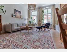 Helles Wohnzimmer mit einem großen, gemusterten Teppich, einer grauen Couch, einem Holztisch und einem Schaukelstuhl mit Hocker. Im Hintergrund befinden sich große Fenster mit Gardinen, Zimmerpflanzen und eine minimalistische Wanddekoration.