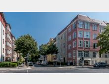 Straßenecke in einer Wohngegend mit mehrstöckigen Altbauten in Pastellfarben, darunter rosa und beige Fassaden mit großen Fenstern. Am Straßenrand stehen Bäume und parkende Autos, während im Vordergrund eine Ampel und ein Fußgängerüberweg zu sehen sind.