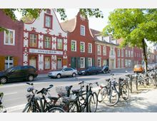 Straßenszene mit roten Backsteinhäusern im historischen Stil, teils mit verziertem Putz, Fenstern und Giebeln. Im Vordergrund stehen geparkte Fahrräder, dahinter geparkte Autos entlang der Straße, gesäumt von Bäumen.