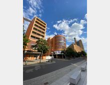 Straßenansicht eines städtischen Bereichs mit modernen Backsteingebäuden. Im Vordergrund stehen Bäume und ein gepflasterter Gehweg, mit einem runden, markanten Gebäude, das mit „Blue Man Group“ beschriftet ist. Der Himmel ist klar mit vereinzelten Wolken.