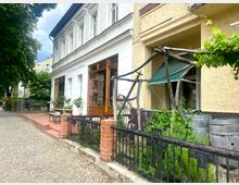 Reihenhausfassade mit großen Fenstern, in denen ein Geschäft mit der Aufschrift „Freude schenken täglich“ erkennbar ist. Im Vordergrund ein Gehweg mit Kopfsteinpflaster, ein kleiner Vorgarten mit Pflanzen, Holzfässern und einem Rankgerüst aus Holz.